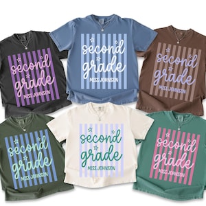 May include: Six short-sleeve t-shirts in various colors including black, blue, brown, olive green, cream, and teal. Each shirt features a central graphic with vertical stripes, the text "second grade" in script, and "MISS JOHNSON".