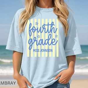 May include: A light blue t-shirt featuring a graphic design with vertical yellow stripes and blue text that reads "fourth grade" and "MISS JOHNSON" with small star accents. The shirt has a crew neckline and short sleeves.