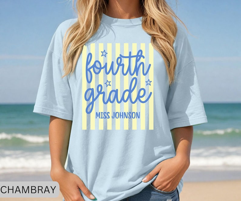 May include: A light blue t-shirt featuring a graphic design with vertical yellow stripes and blue text that reads "fourth grade" and "MISS JOHNSON" with small star accents. The shirt has a crew neckline and short sleeves.