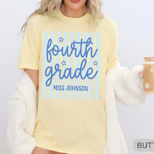 May include: A pale yellow short-sleeve t-shirt featuring a light blue vertical striped rectangle graphic. The text reads fourth grade in a blue script font with small stars, and MISS JOHNSON in blue block letters below the script text.