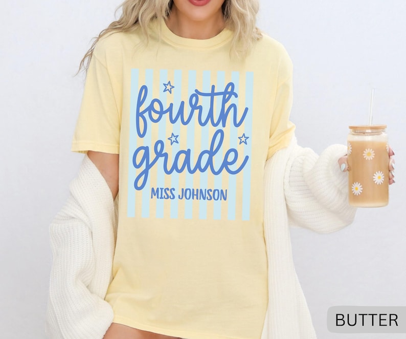 May include: A pale yellow short-sleeve t-shirt featuring a light blue vertical striped rectangle graphic. The text reads fourth grade in a blue script font with small stars, and MISS JOHNSON in blue block letters below the script text.