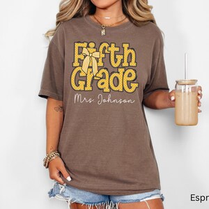 K&ouml;nnte beinhalten: Ein espressofarbenes Kurzarm-T-Shirt mit dem gelben Schriftzug Fifth Grade, einer gelben Schleifengrafik und dem wei&szlig;en Schreibschrift-Text Mrs. Johnson auf der Vorderseite. Das T-Shirt hat einen Rundhalsausschnitt und eine lockere Passform.