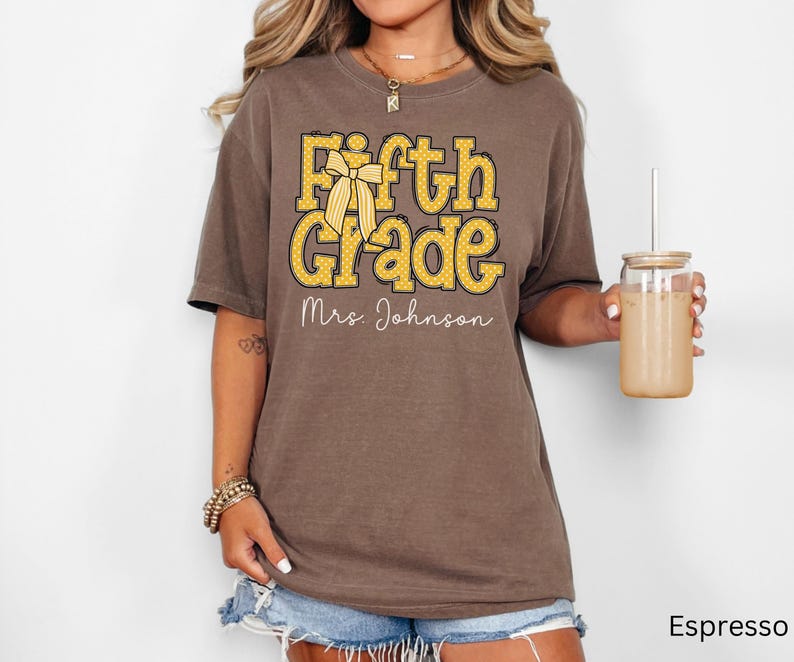 K&ouml;nnte beinhalten: Ein espressofarbenes Kurzarm-T-Shirt mit dem gelben Schriftzug Fifth Grade, einer gelben Schleifengrafik und dem wei&szlig;en Schreibschrift-Text Mrs. Johnson auf der Vorderseite. Das T-Shirt hat einen Rundhalsausschnitt und eine lockere Passform.