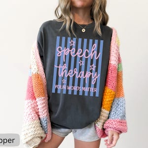 May include: A charcoal gray t-shirt featuring a light blue vertical striped rectangle with pink cursive text that reads speech therapy and block text that reads YOUR WORDS MATTER, accented by small pink stars, worn under a multicolored chunky knit cardigan.