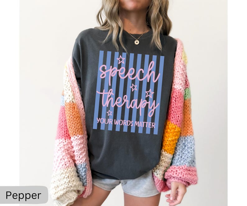 May include: A charcoal gray t-shirt featuring a light blue vertical striped rectangle with pink cursive text that reads speech therapy and block text that reads YOUR WORDS MATTER, accented by small pink stars, worn under a multicolored chunky knit cardigan.