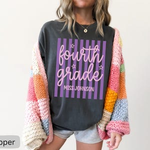 May include: A charcoal gray t-shirt featuring a graphic with vertical purple stripes and the text "fourth grade" in pink cursive script, accented by small stars and the name "MISS JOHNSON" in white block letters, worn under a chunky knit cardigan.