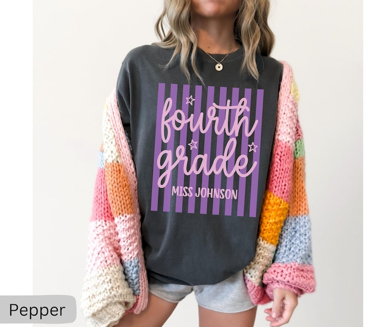 May include: A charcoal gray t-shirt featuring a graphic with vertical purple stripes and the text "fourth grade" in pink cursive script, accented by small stars and the name "MISS JOHNSON" in white block letters, worn under a chunky knit cardigan.
