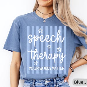 May include: A blue jean colored short-sleeve t-shirt featuring a white graphic design with vertical stripes, stars, and the text "speech therapy" in a cursive font above the block text "YOUR WORDS MATTER" centered on the front of the garment.