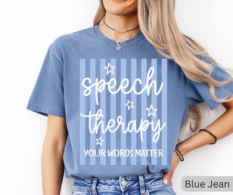 May include: A blue jean colored short-sleeve t-shirt featuring a white graphic design with vertical stripes, stars, and the text "speech therapy" in a cursive font above the block text "YOUR WORDS MATTER" centered on the front of the garment.