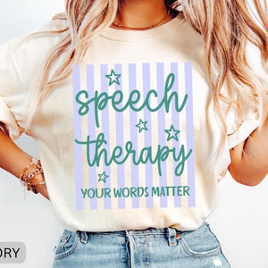 May include: An ivory short-sleeve t-shirt featuring a graphic design with vertical light purple stripes. The text reads "speech therapy" in a dark green script font, accompanied by four small stars and the text "YOUR WORDS MATTER" below.