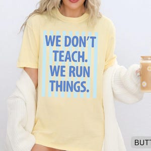 May include: A light yellow t-shirt with the text "WE DON'T TEACH. WE RUN THINGS." in blue. The text is centered within a light blue striped rectangle. A glass with daisy decals and a straw is held in the hand.