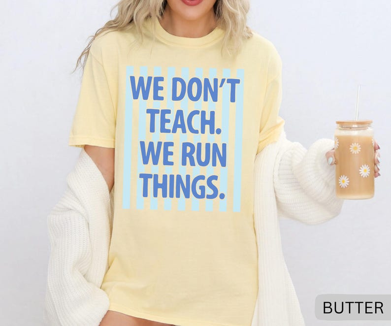 May include: A light yellow t-shirt with the text "WE DON'T TEACH. WE RUN THINGS." in blue. The text is centered within a light blue striped rectangle. A glass with daisy decals and a straw is held in the hand.