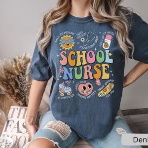 Pode incluir: Uma camiseta azul escura com um design gr&aacute;fico colorido que diz "School Nurse" com v&aacute;rias ilustra&ccedil;&otilde;es e texto ao redor. O texto inclui "Smile, it's medicine", "Quick checkup", "Wiggle your fingers and toes", "Bright bandage for you", e mais.