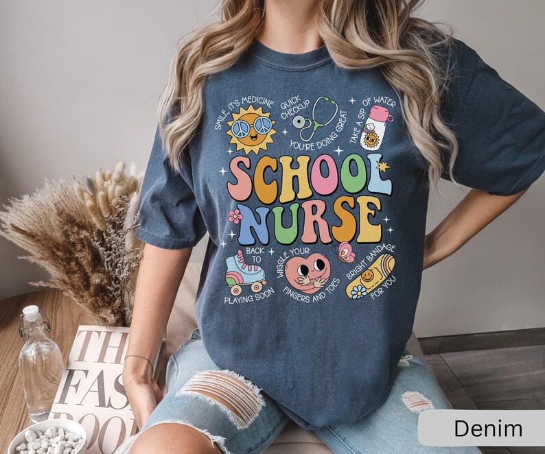 Pode incluir: Uma camiseta azul escura com um design gr&aacute;fico colorido que diz "School Nurse" com v&aacute;rias ilustra&ccedil;&otilde;es e texto ao redor. O texto inclui "Smile, it's medicine", "Quick checkup", "Wiggle your fingers and toes", "Bright bandage for you", e mais.