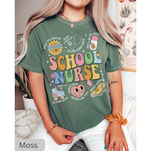 Pode incluir: Camiseta verde de manga curta com uma estampa gr&aacute;fica colorida que diz "School Nurse" e inclui v&aacute;rias ilustra&ccedil;&otilde;es de materiais escolares e artigos m&eacute;dicos. O texto na camiseta diz: "Smile it's medicine, Quick checkup, You're doing great, Take a sip of water, Wiggle your fingers and toes, Bright bandage for you, Back to playing soon."