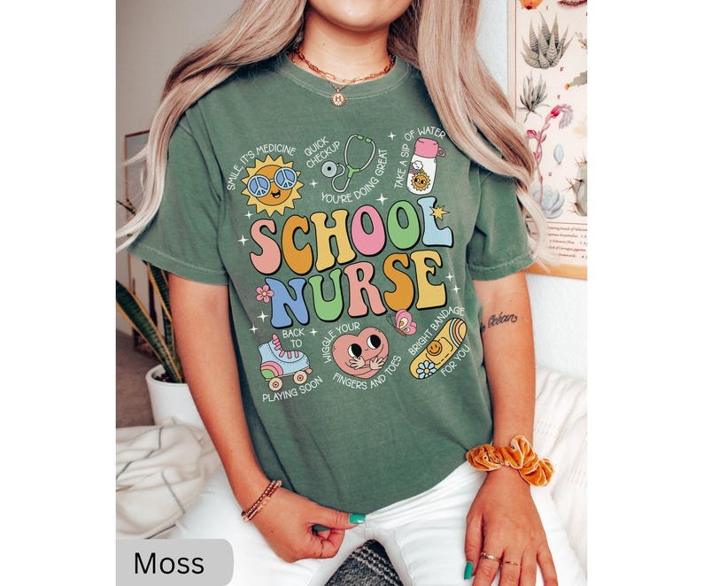 Pode incluir: Camiseta verde de manga curta com uma estampa gr&aacute;fica colorida que diz "School Nurse" e inclui v&aacute;rias ilustra&ccedil;&otilde;es de materiais escolares e artigos m&eacute;dicos. O texto na camiseta diz: "Smile it's medicine, Quick checkup, You're doing great, Take a sip of water, Wiggle your fingers and toes, Bright bandage for you, Back to playing soon."