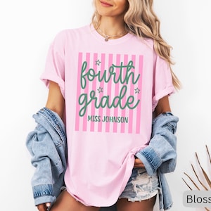 May include: A light pink t-shirt featuring a vertical pink striped rectangle graphic. The text "fourth grade" is printed in a green cursive font, accompanied by small green stars. Below, "MISS JOHNSON" is printed in a green sans-serif font.
