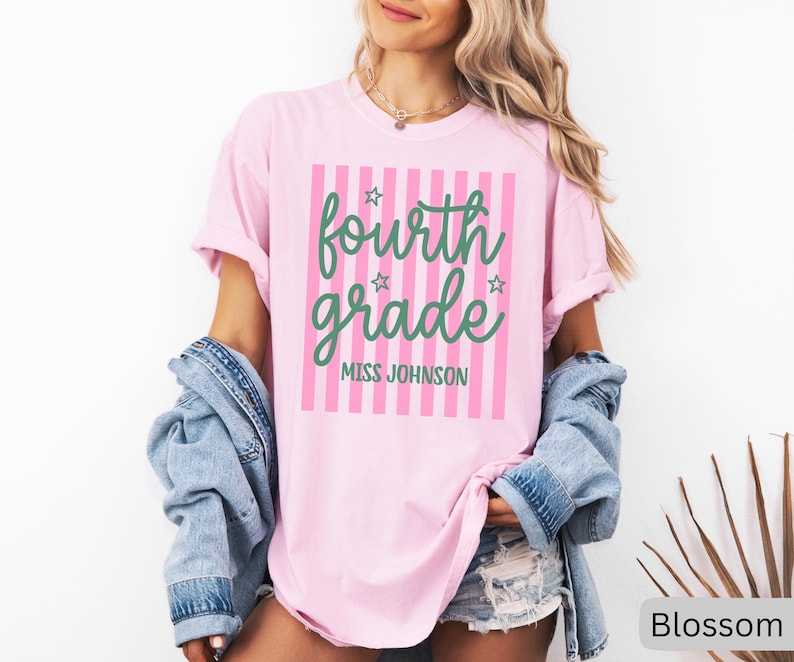 May include: A light pink t-shirt featuring a vertical pink striped rectangle graphic. The text "fourth grade" is printed in a green cursive font, accompanied by small green stars. Below, "MISS JOHNSON" is printed in a green sans-serif font.