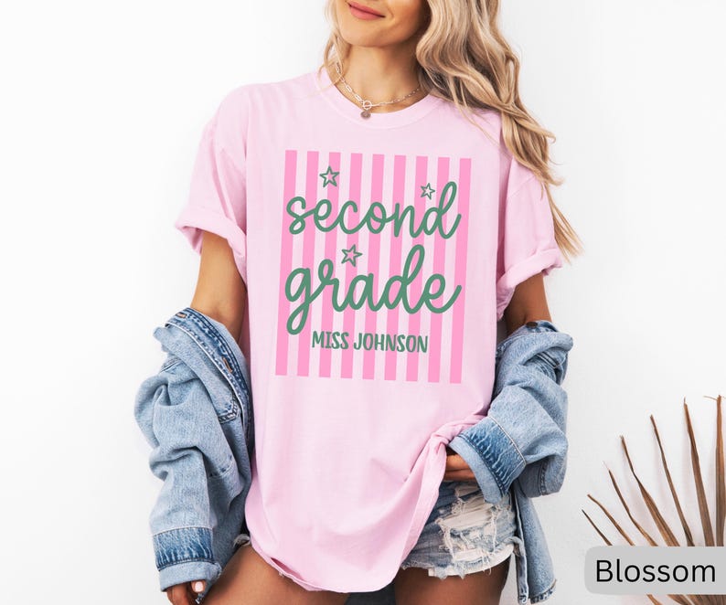 May include: A light pink short-sleeve t-shirt featuring a vertical pink striped rectangle with the green text second grade and MISS JOHNSON, accented by three small green star outlines. The shirt is worn with a blue denim jacket and denim shorts.