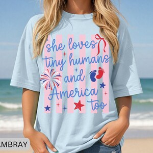 May include: A light blue t-shirt featuring a graphic with pink vertical stripes and the text "she loves tiny humans and America too". The design includes a red bow, a firework, blue and red baby footprints, and several red and blue stars.