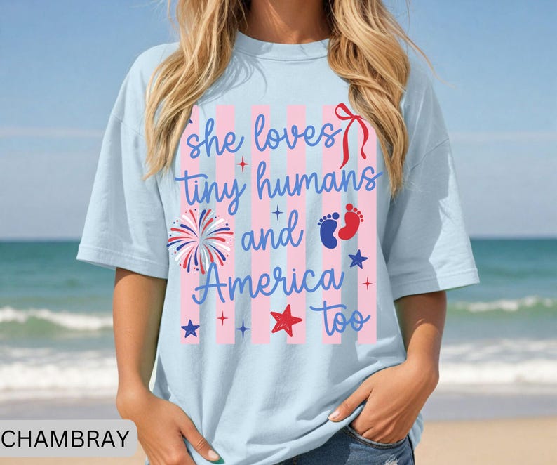 May include: A light blue t-shirt featuring a graphic with pink vertical stripes and the text "she loves tiny humans and America too". The design includes a red bow, a firework, blue and red baby footprints, and several red and blue stars.