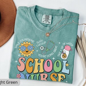 Pode incluir: Camiseta verde claro com um gr&aacute;fico de estilo retro que diz "School Nurse" com um sol sorridente, um estetosc&oacute;pio e uma garrafa de &aacute;gua. A camiseta tem um visual desgastado e &eacute; feita de um tecido macio e confort&aacute;vel.