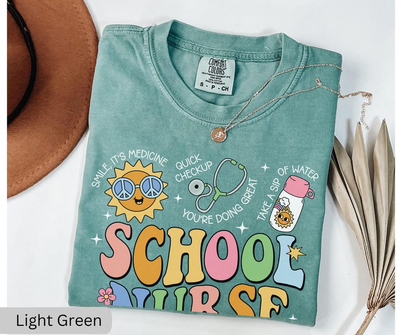 Pode incluir: Camiseta verde claro com um gr&aacute;fico de estilo retro que diz "School Nurse" com um sol sorridente, um estetosc&oacute;pio e uma garrafa de &aacute;gua. A camiseta tem um visual desgastado e &eacute; feita de um tecido macio e confort&aacute;vel.