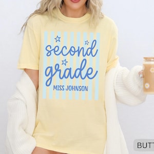 May include: A pale yellow short-sleeve t-shirt featuring a light blue vertical striped rectangle with the blue text "second grade" in a cursive font, three small star outlines, and the block text "MISS JOHNSON" printed underneath.