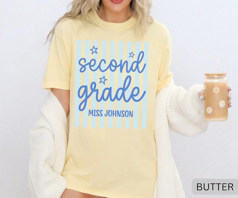 May include: A pale yellow short-sleeve t-shirt featuring a light blue vertical striped rectangle with the blue text "second grade" in a cursive font, three small star outlines, and the block text "MISS JOHNSON" printed underneath.