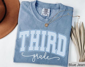 Könnte beinhalten: Ein blaues Comfort Colors T-Shirt mit den Worten "THIRD grade" in einem weiß-blau gestreiften Design. Eine goldene Halskette mit Anhänger liegt über dem Shirt. Das Shirt ist gefaltet.