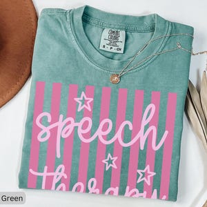 May include: A light green short-sleeve t-shirt with a crew neckline and a Comfort Colors brand label. The front features a graphic with vertical pink stripes, three outlined stars, and the text "speech therapy" written in a light pink script font.