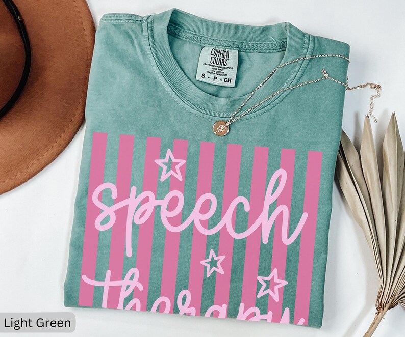 May include: A light green short-sleeve t-shirt with a crew neckline and a Comfort Colors brand label. The front features a graphic with vertical pink stripes, three outlined stars, and the text "speech therapy" written in a light pink script font.