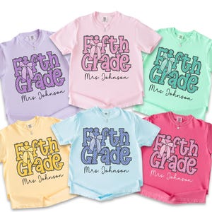 K&ouml;nnte beinhalten: Sechs kurz&auml;rmelige T-Shirts in Lavendel, Hellrosa, Mintgr&uuml;n, Blassgelb, Hellblau und leuchtendem Pink. Jedes T-Shirt zeigt den Text Fifth Grade in einem Punktemuster mit einer Schleifengrafik und dem darunter gedruckten Namen Mrs. Johnson.