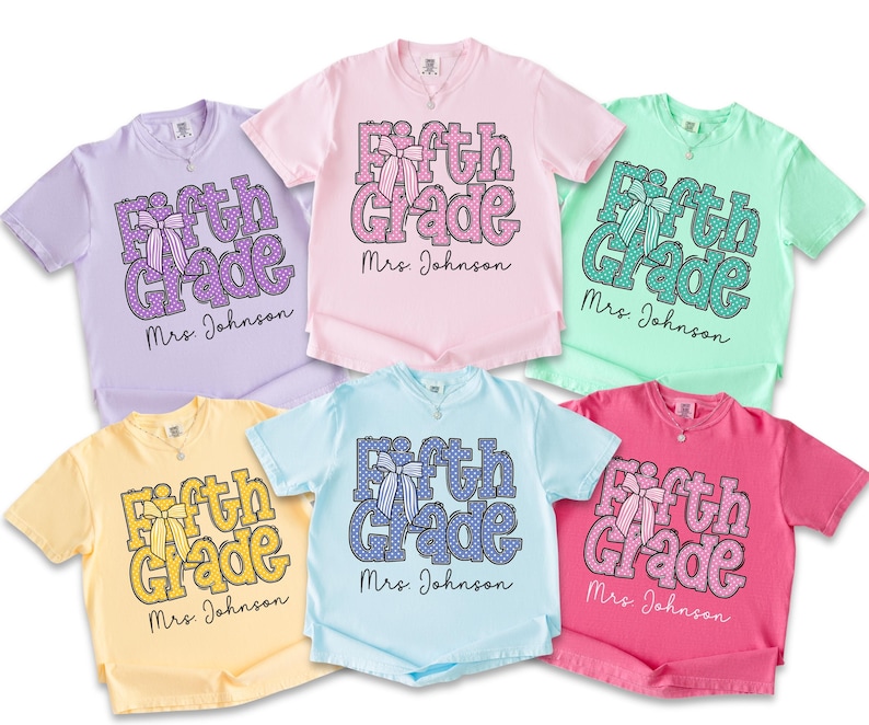 K&ouml;nnte beinhalten: Sechs kurz&auml;rmelige T-Shirts in Lavendel, Hellrosa, Mintgr&uuml;n, Blassgelb, Hellblau und leuchtendem Pink. Jedes T-Shirt zeigt den Text Fifth Grade in einem Punktemuster mit einer Schleifengrafik und dem darunter gedruckten Namen Mrs. Johnson.