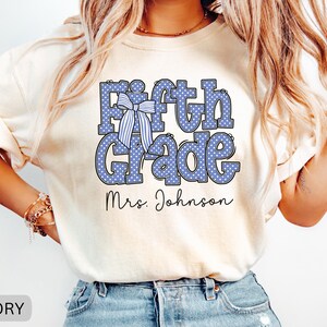 K&ouml;nnte beinhalten: Ein elfenbeinfarbenes T-Shirt mit dem Text Fifth Grade in Blau mit wei&szlig;en Punkten. Eine blau-wei&szlig; gestreifte Schleife ist &uuml;ber dem Buchstaben F platziert. Unter dem Text ist der Name Mrs. Johnson in einer schwarzen Schreibschrift gedruckt.