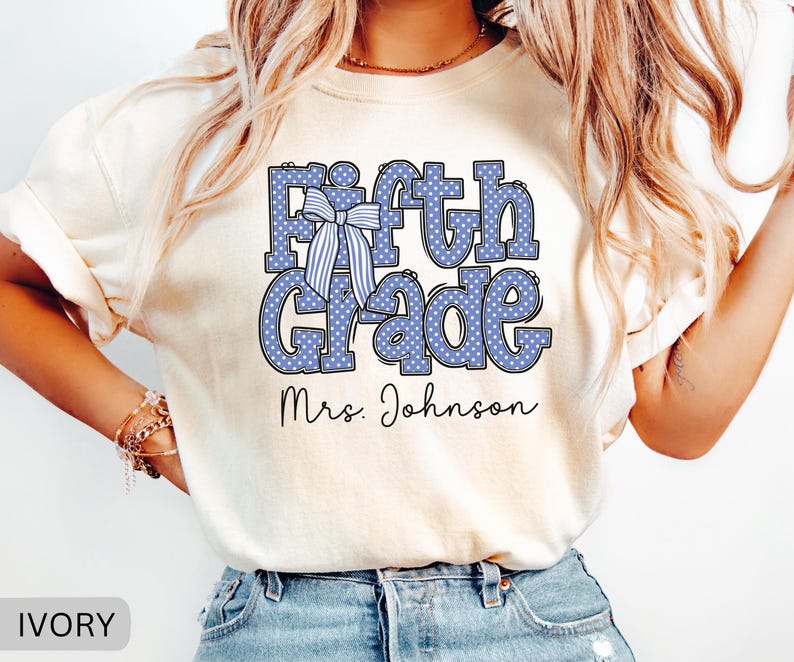 K&ouml;nnte beinhalten: Ein elfenbeinfarbenes T-Shirt mit dem Text Fifth Grade in Blau mit wei&szlig;en Punkten. Eine blau-wei&szlig; gestreifte Schleife ist &uuml;ber dem Buchstaben F platziert. Unter dem Text ist der Name Mrs. Johnson in einer schwarzen Schreibschrift gedruckt.
