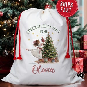 May include: White Christmas gift bag with red drawstring closure. The bag features a ballerina with a Christmas tree and the text "Special Delivery For Olivia". The bag is surrounded by Christmas decorations.