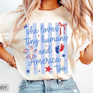 May include: An ivory t-shirt featuring a blue striped background with the text "she loves tiny humans and America too" in blue script. The design includes a red bow, a firework graphic, red and blue baby footprints, and various stars.