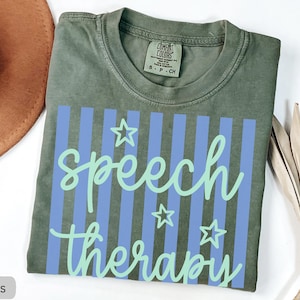 May include: A moss green short-sleeved t-shirt featuring a graphic print of vertical periwinkle blue stripes. The text "speech therapy" is written in a light mint green cursive font, accompanied by three outlined star shapes in the same color.