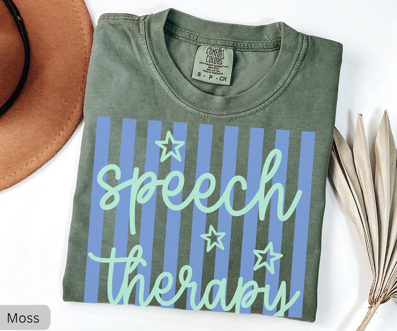May include: A moss green short-sleeved t-shirt featuring a graphic print of vertical periwinkle blue stripes. The text "speech therapy" is written in a light mint green cursive font, accompanied by three outlined star shapes in the same color.