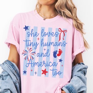 May include: A light pink t-shirt featuring a blue and white vertical striped background with the text 'she loves tiny humans and America too' in blue script. The design includes a red bow, a firework, blue and red baby footprints, and stars.