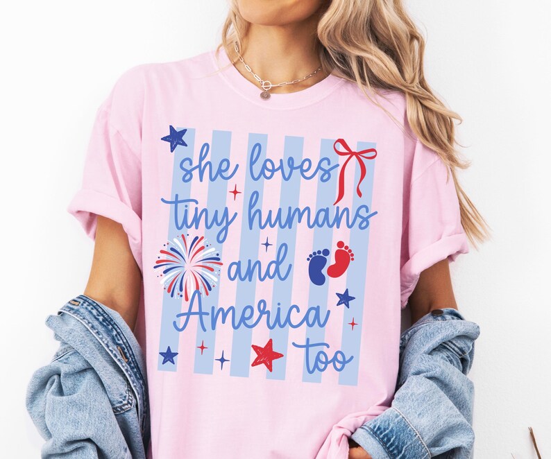May include: A light pink t-shirt featuring a blue and white vertical striped background with the text 'she loves tiny humans and America too' in blue script. The design includes a red bow, a firework, blue and red baby footprints, and stars.