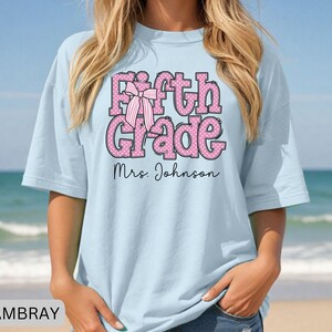 K&ouml;nnte beinhalten: Ein hellblaues T-Shirt mit dem Text Fifth Grade in Pink mit wei&szlig;en Punkten und einer Grafik einer gestreiften rosa Schleife. Unter dem Text ist der Name Mrs. Johnson in einer schwarzen Schreibschrift gedruckt. Das Shirt hat einen Rundhalsausschnitt.