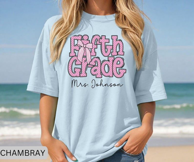 K&ouml;nnte beinhalten: Ein hellblaues T-Shirt mit dem Text Fifth Grade in Pink mit wei&szlig;en Punkten und einer Grafik einer gestreiften rosa Schleife. Unter dem Text ist der Name Mrs. Johnson in einer schwarzen Schreibschrift gedruckt. Das Shirt hat einen Rundhalsausschnitt.