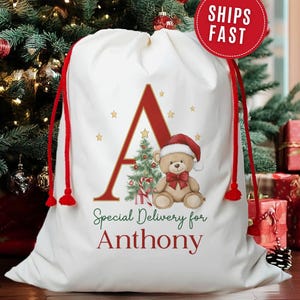 May include: A white Christmas gift bag with red drawstrings and a festive design. The bag features a large red letter "A", a teddy bear wearing a Santa hat, and the text "Special Delivery for Anthony". Gold stars and a Christmas tree are also included.
