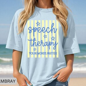 May include: A light blue t-shirt featuring a central graphic with vertical yellow and white stripes. The text reads speech therapy in blue script, accompanied by four blue star outlines and the text YOUR WORDS MATTER in blue block letters.