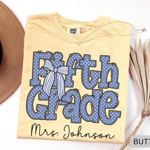 K&ouml;nnte beinhalten: Ein buttergelbes T-Shirt mit dem Text Fifth Grade in Blau mit wei&szlig;en Punkten und einer blau-wei&szlig; gestreiften Schleifengrafik. Unter dem Text ist der Name Mrs. Johnson in schwarzer Schreibschrift gedruckt. Das T-Shirt hat ein Comfort Colors Etikett.