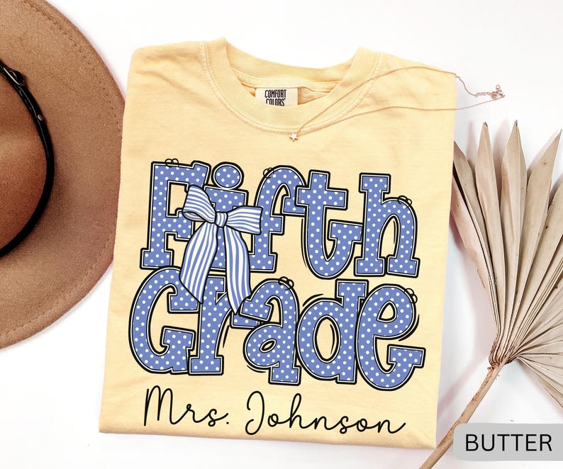 K&ouml;nnte beinhalten: Ein buttergelbes T-Shirt mit dem Text Fifth Grade in Blau mit wei&szlig;en Punkten und einer blau-wei&szlig; gestreiften Schleifengrafik. Unter dem Text ist der Name Mrs. Johnson in schwarzer Schreibschrift gedruckt. Das T-Shirt hat ein Comfort Colors Etikett.