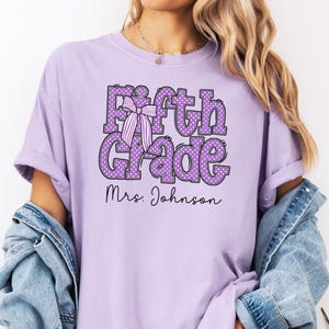 K&ouml;nnte beinhalten: Ein hellviolettes Kurzarm-T-Shirt mit dem Text Fifth Grade in gro&szlig;en, lila gepunkteten Buchstaben und einer gestreiften Schleifengrafik. Unter dem Haupttext ist der Name Mrs. Johnson in einer schwarzen Schreibschrift aufgedruckt.