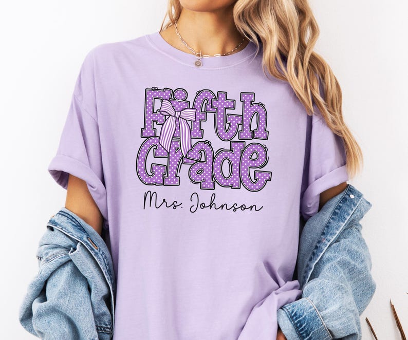 K&ouml;nnte beinhalten: Ein hellviolettes Kurzarm-T-Shirt mit dem Text Fifth Grade in gro&szlig;en, lila gepunkteten Buchstaben und einer gestreiften Schleifengrafik. Unter dem Haupttext ist der Name Mrs. Johnson in einer schwarzen Schreibschrift aufgedruckt.